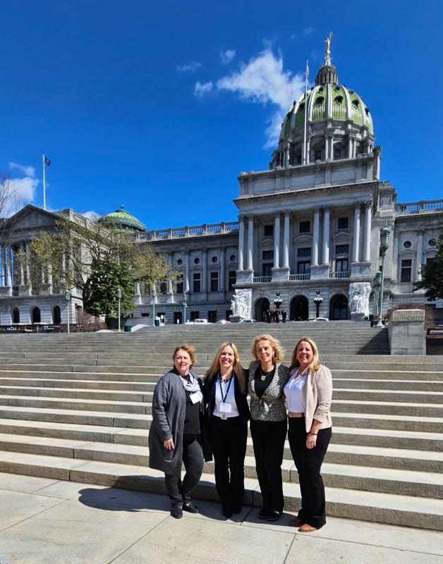  Four people smile for a photo outside in front of the Pennsylvania State Capitol Complex